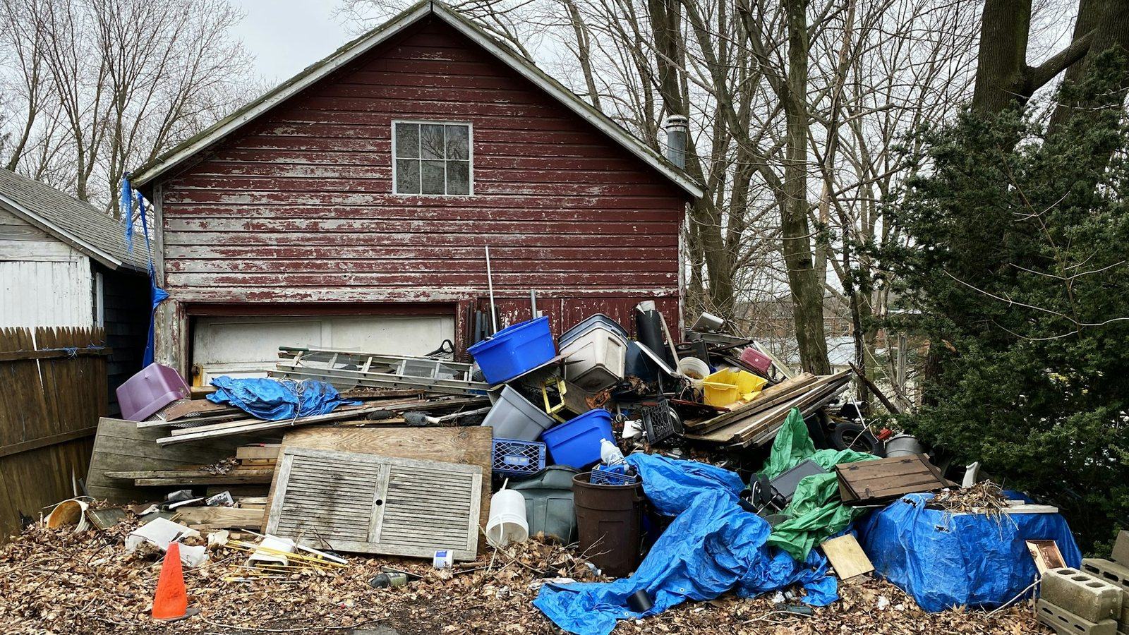 A head-high pile of scrap timber, bins, tarps and broken furniture heaped against the side of a red outbuilding