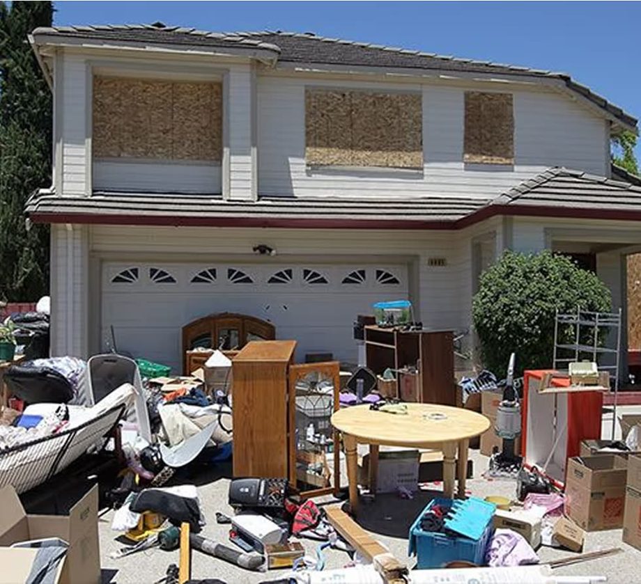 A boarded-up two-storey house with cabinets, tables, boxes and tools from an estate cleanout spread across the driveway